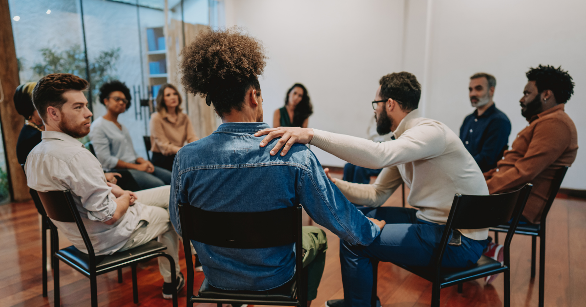 Diverse group of people in a support circle, one man comforting another with hand on shoulder.