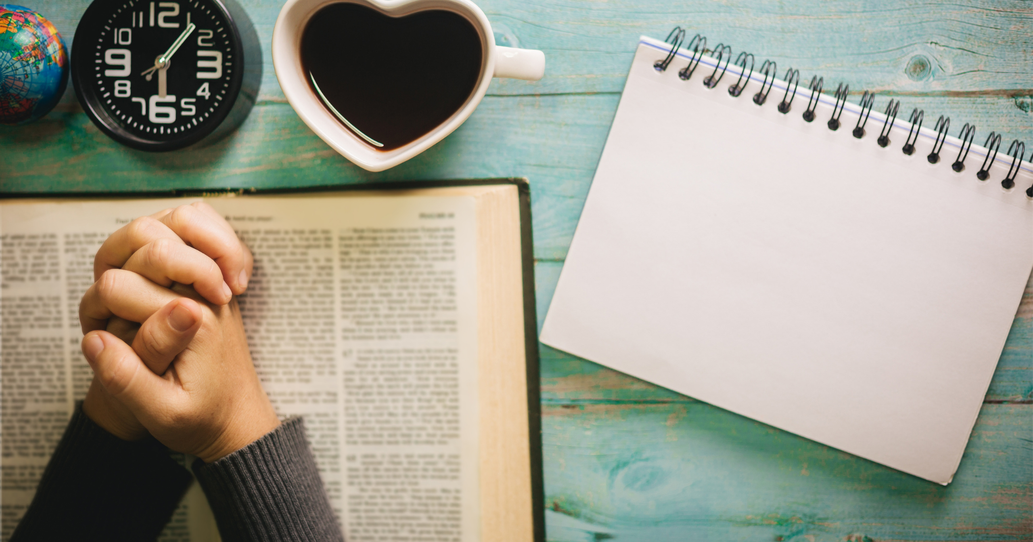 A person with folded hands in prayer over an open Bible, next to a notebook, coffee, and clock, symbolizing daily devotion and personal Bible study.