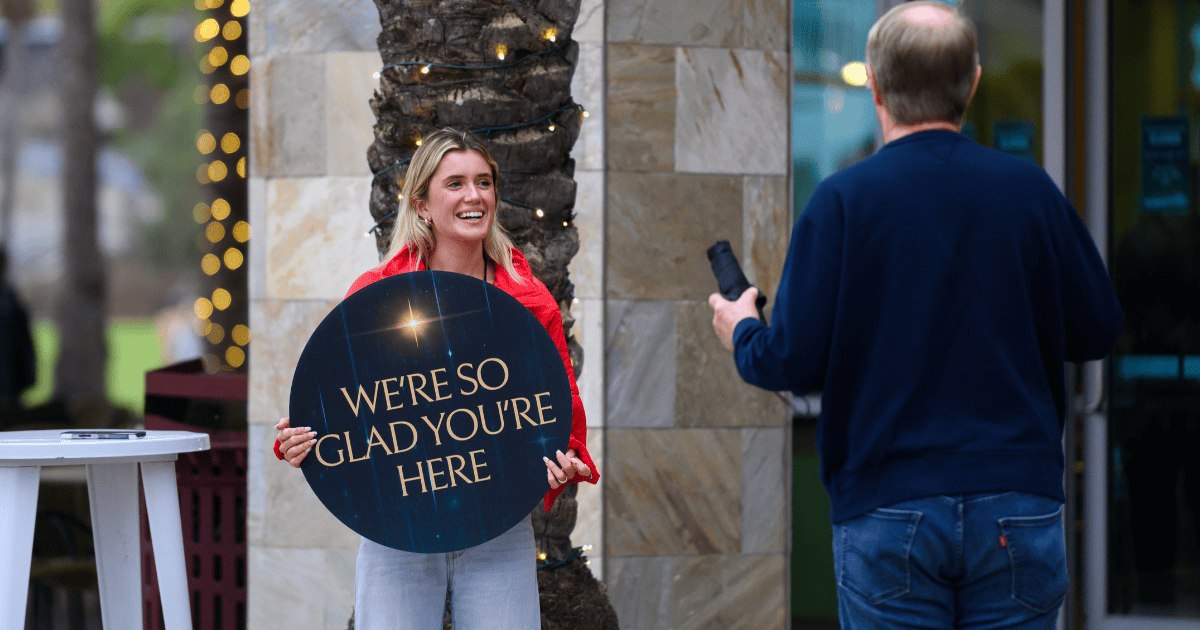 A smiling church volunteer in a red jacket holding a "We're so glad you're here" sign, representing the strategic winter window for inviting people into a new year discipleship launch.